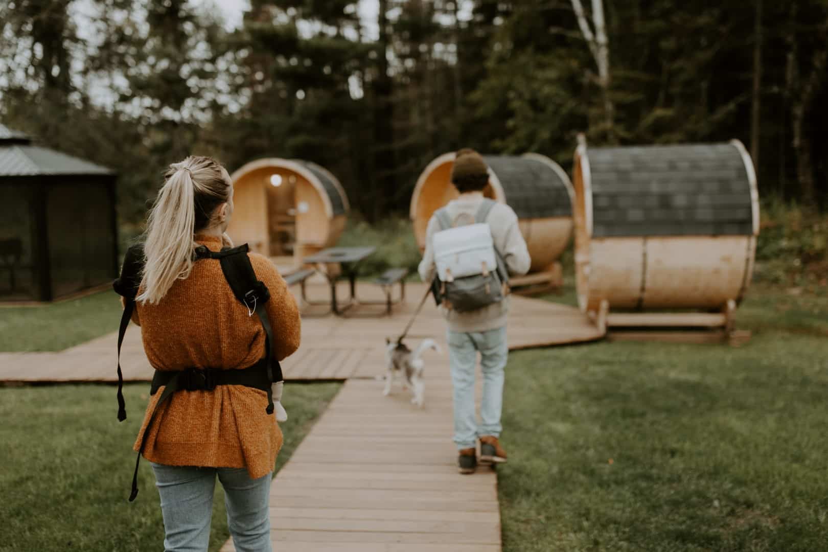 Couple walking dog near barrel-shaped cabins in forest.