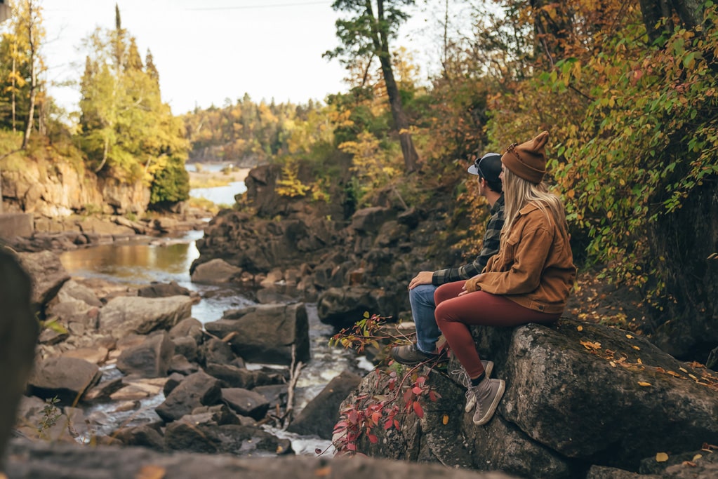 Two hikers resting by forested riverbank in autumn.