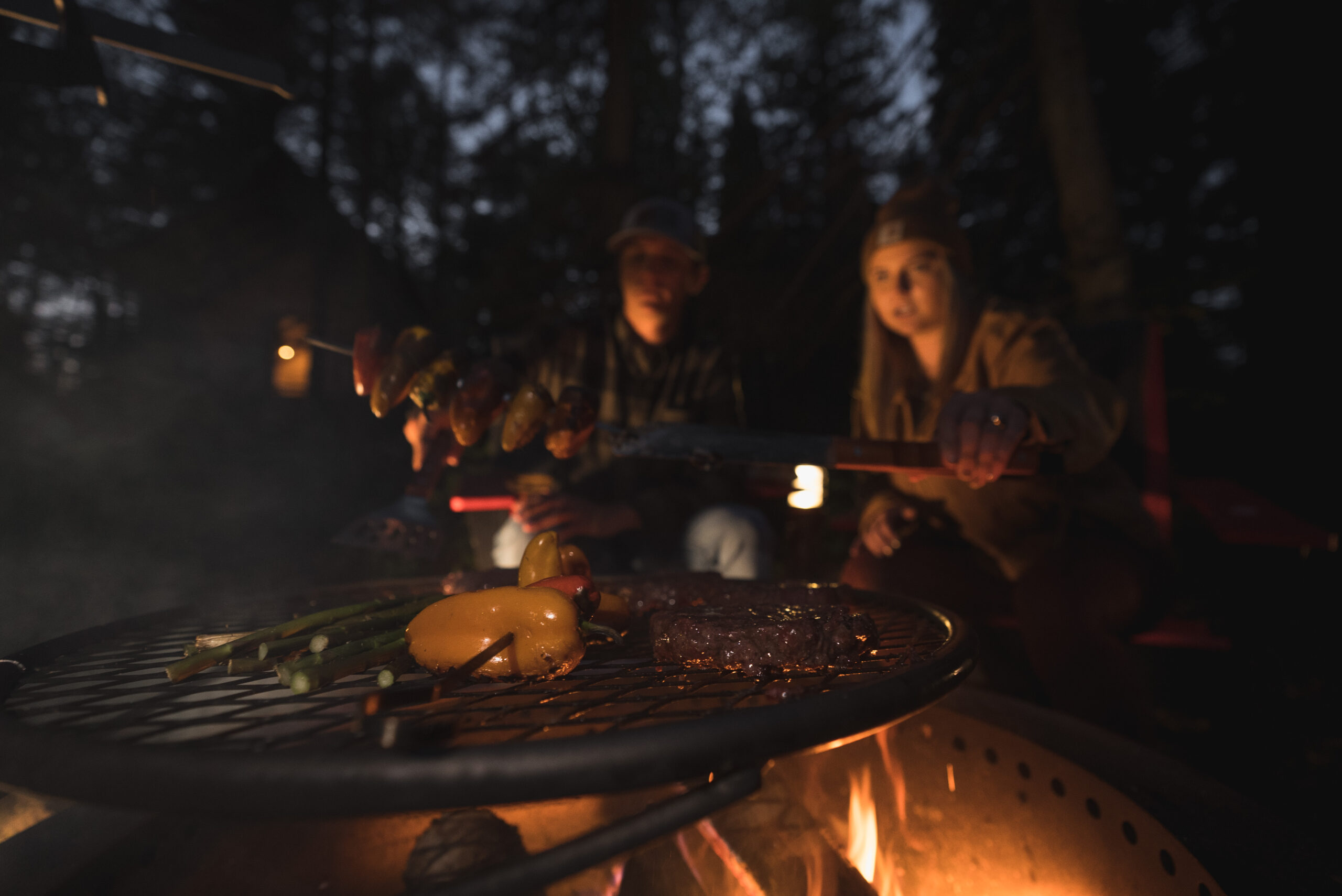 People grilling vegetables and meat at campfire.