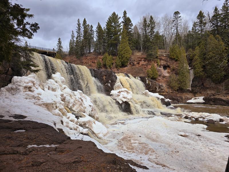 Waterfall flowing over snowy rocks with pine trees.