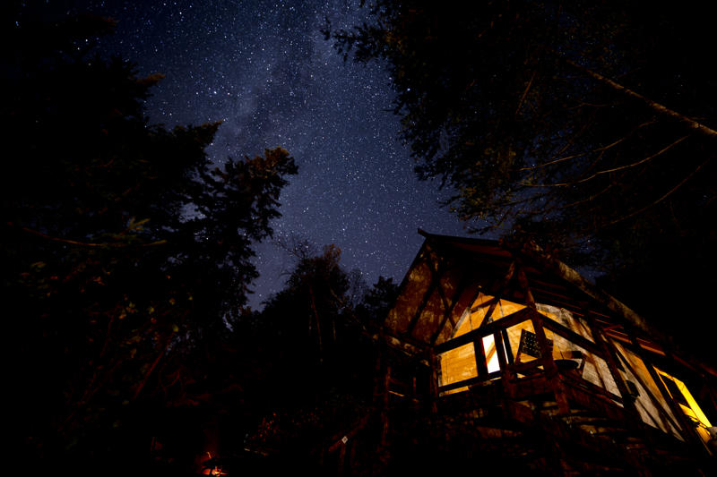 Starlit night sky over illuminated cabin in forest.