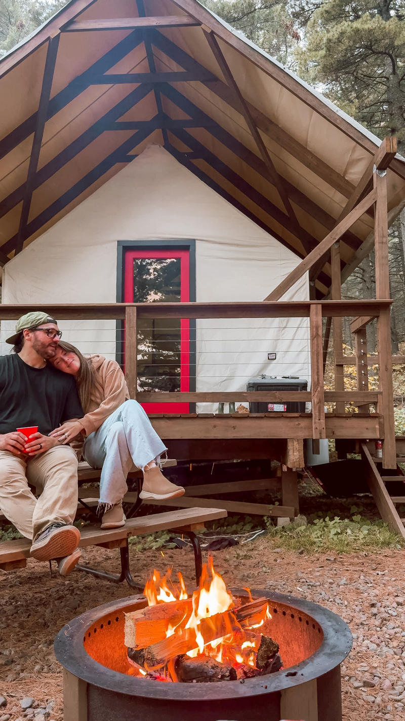 Couple relaxing by campfire outside cabin in woods.