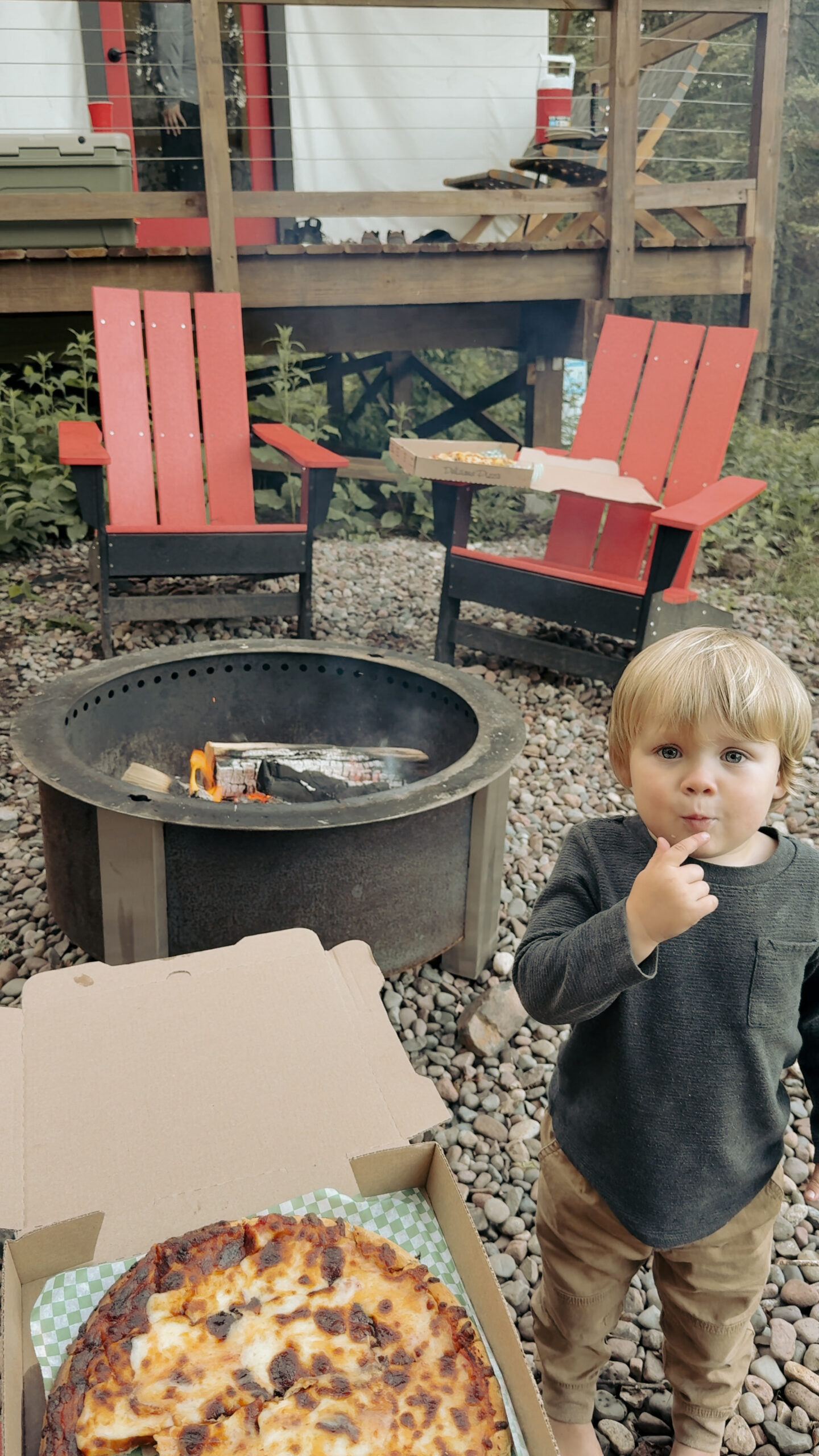 boypizzabyfire Child near pizza box by outdoor fire pit.