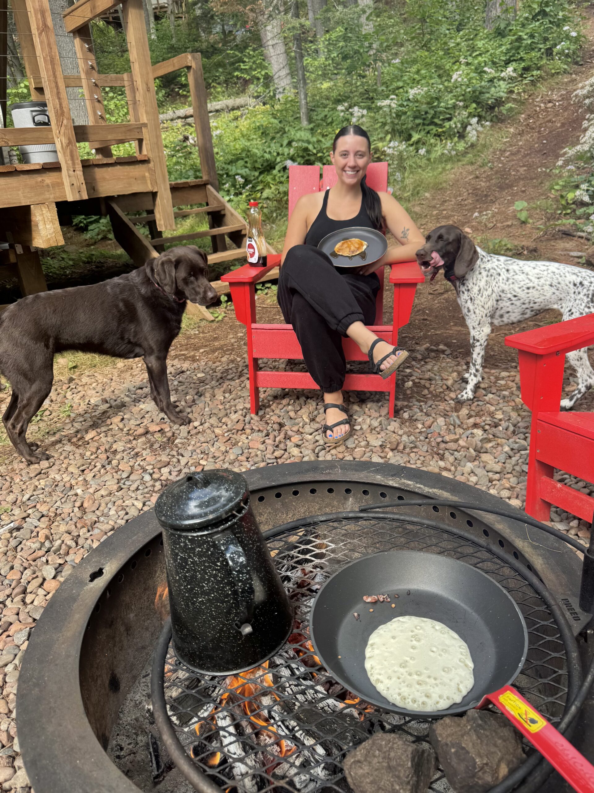 Woman enjoying pancakes with dogs by campfire.