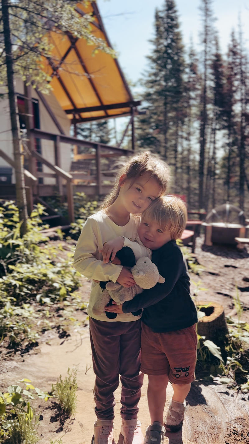 kidsinfrontoftent Two children hug outdoors near cabin.