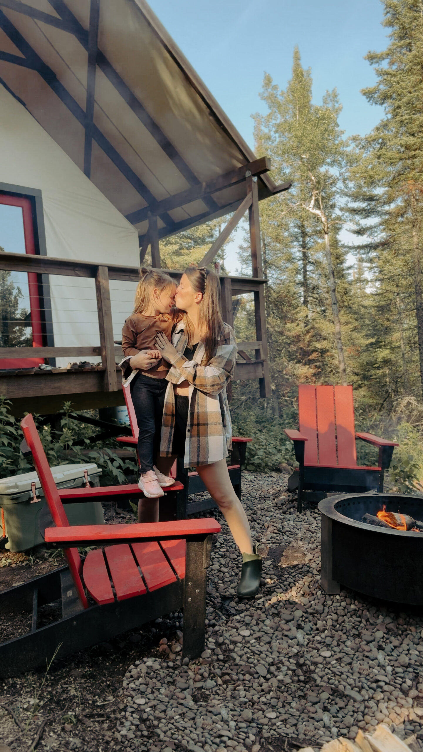 mom&daughterinfrontoftent Mother and child by campfire, wooden cabin background.