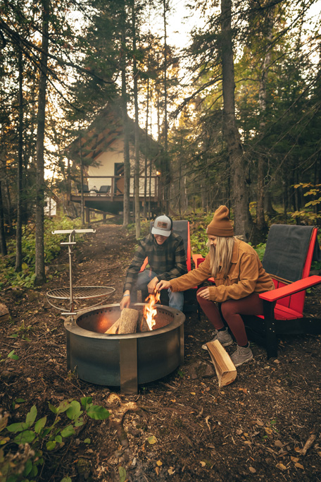 Two people by a campfire near a cabin.