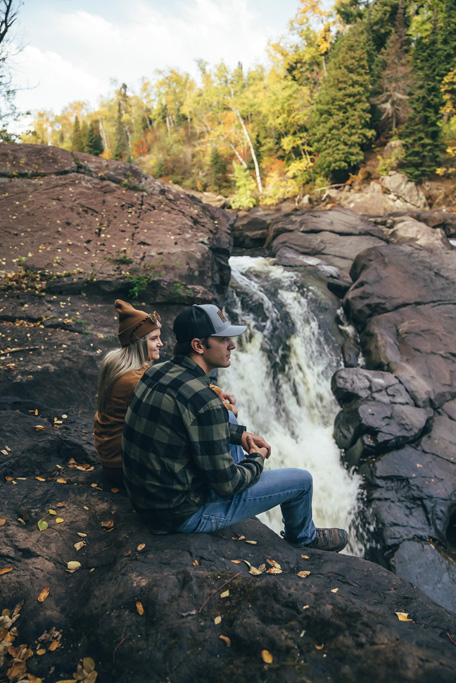 northshorecampingco fall2022 images 70 Couple sitting by waterfall in autumn forest.